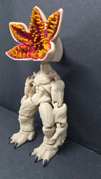 A 3D-printed articulated toy figure of a monster with a beige body, a mouth resembling a clover with yellow and red teeth, and black claws, standing on a dark gray surface.