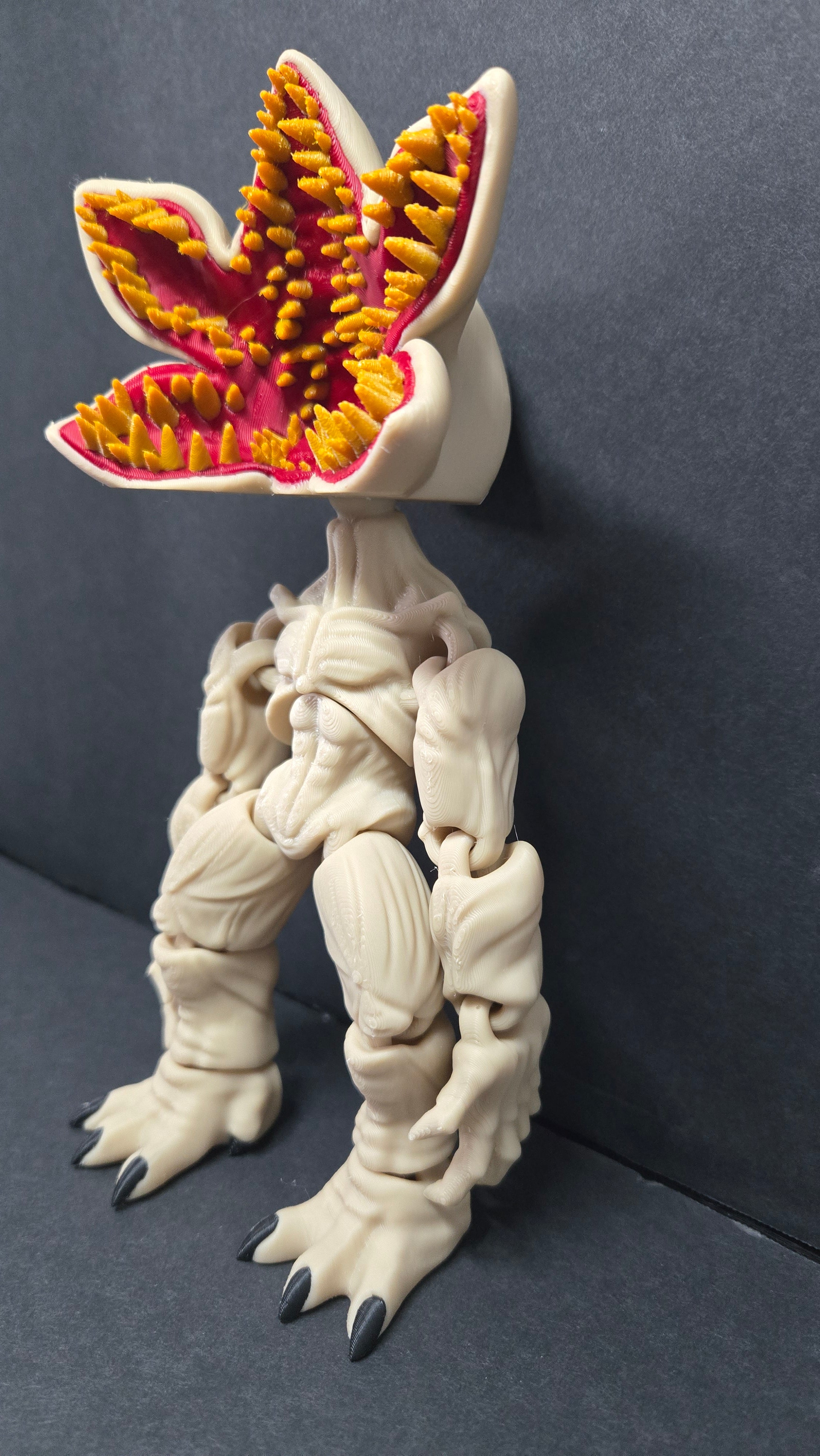 A 3D-printed articulated toy figure of a monster with a beige body, a mouth resembling a clover with yellow and red teeth, and black claws, standing on a dark gray surface.