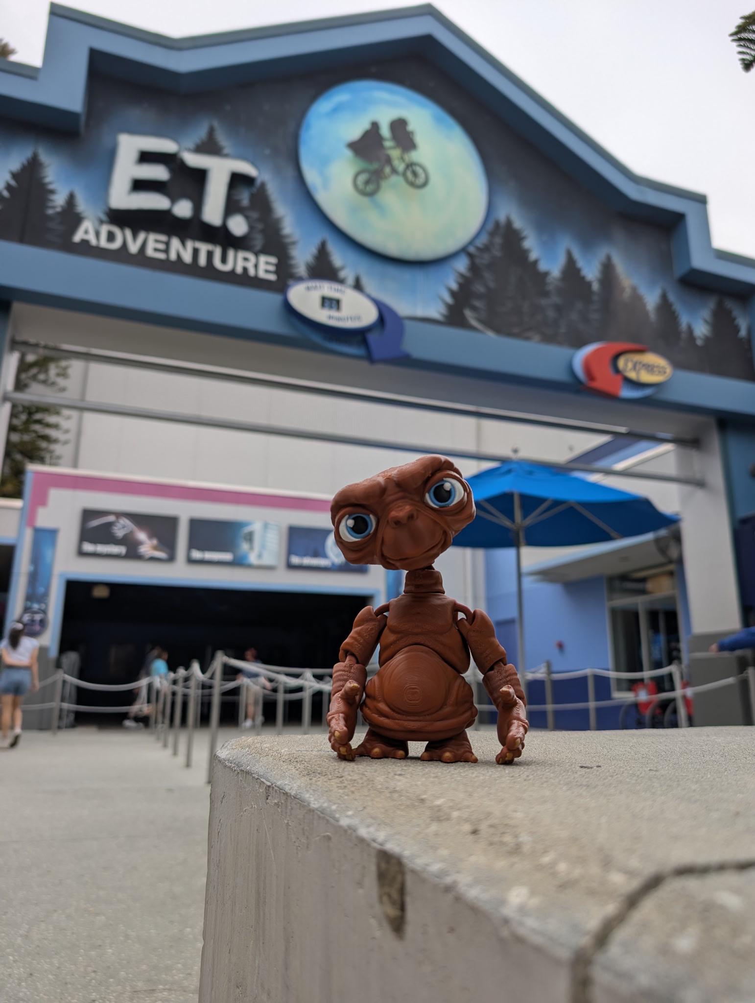 Full-color 3D printed E.T. flexi figure standing upright on a stone ledge in front of the E.T. Adventure theme park entrance