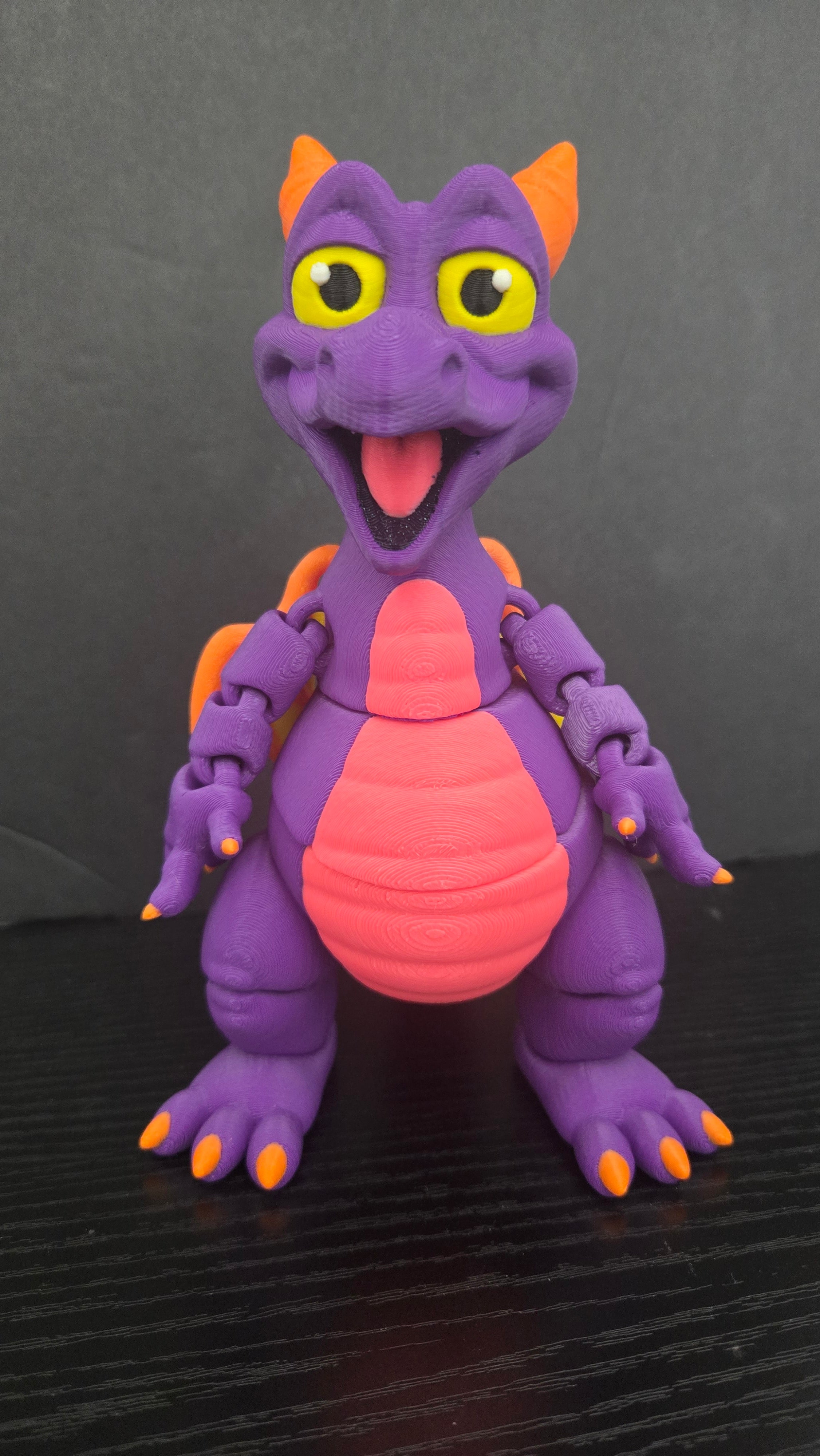 A purple dragon toy figurine with orange horns, claws, and tail, yellow eyes, and a pink belly, standing on a dark wooden surface against a dark gray background.