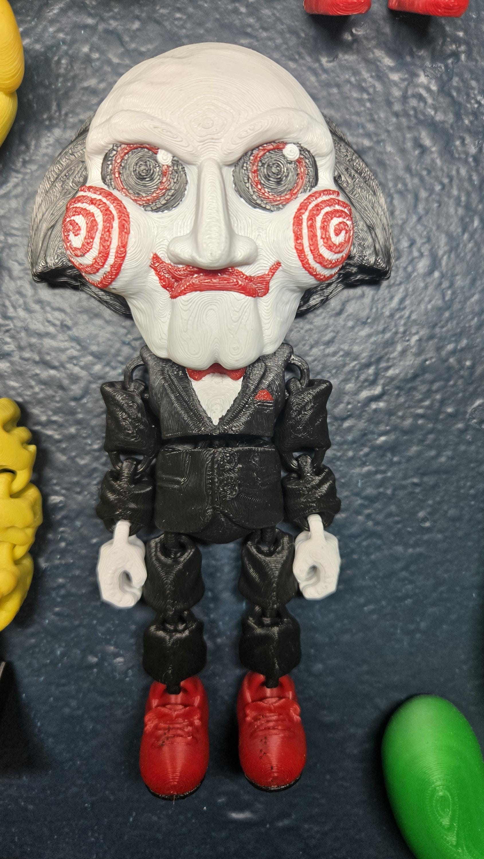 Straight front view focusing on the signature red cheek spirals, black eyes, and bright red bow tie of the flexible Billy the Puppet figure