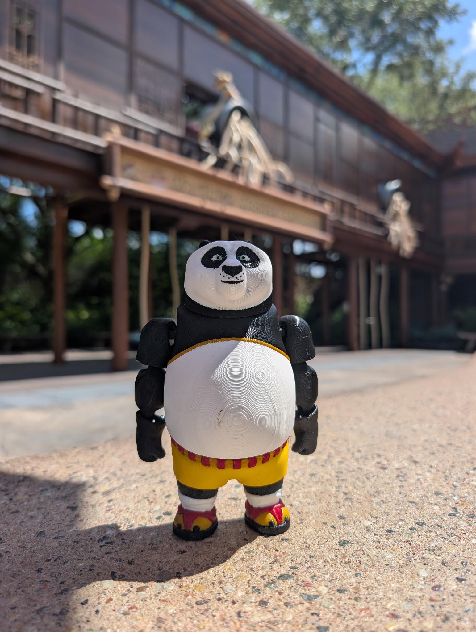 A hand holding up the 3D printed Kung Fu Panda Po flexi figure against a blurred background of Universal Studios theme park attractions