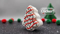 A Christmas tree-shaped white item with red and green decorative details, shown on a dark surface with blurred red and green pom-poms and a small green tree in the background. A 'Clicker!' badge appears on the right.