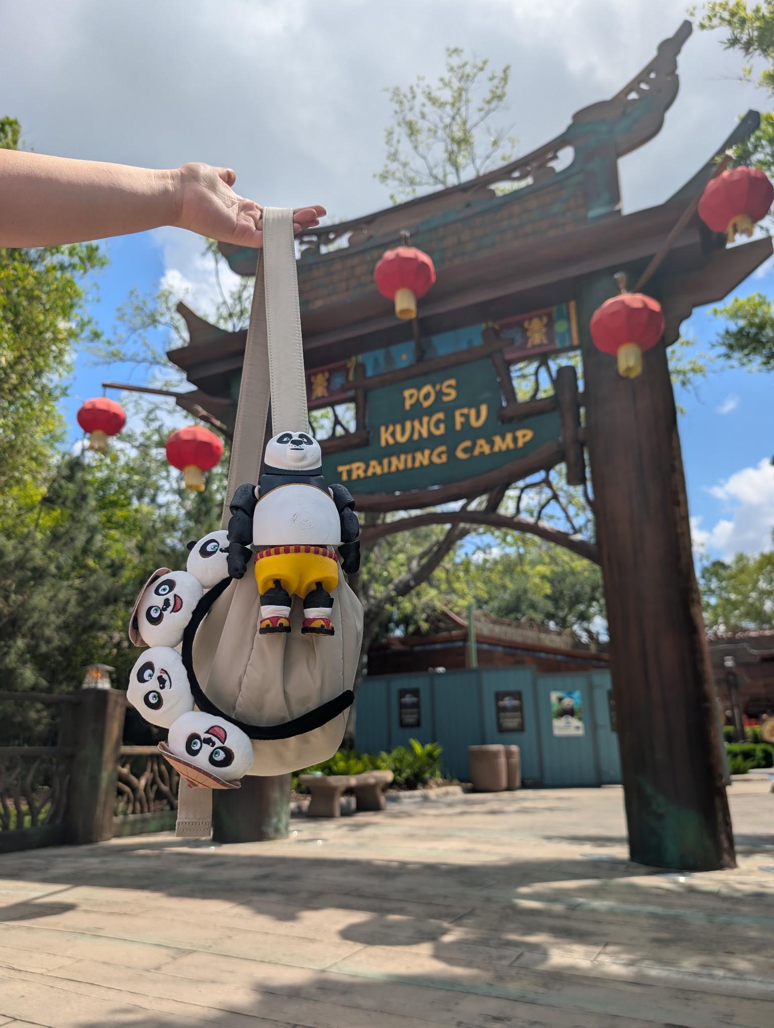 The articulated Kung Fu Panda Po figure being flexed and held up high with a scenic Universal Studios theme park walkway in the background