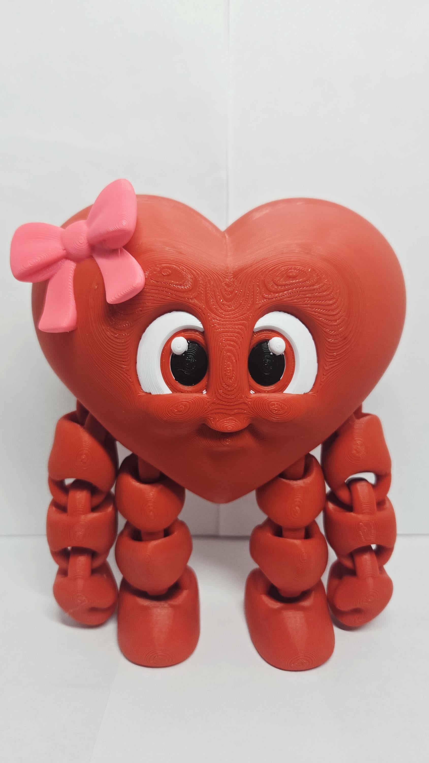 3D printed red Valentine heart figure with a pastel pink bow standing upright on a clean white desk surface